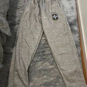 Grå brazil set - Säljer en grå hoodie med dragkedja och huva. Tröjan har ett broderat emblem på bröstet och ribbade muddar vid ärmslut och nederkant. Perfekt för en avslappnad stil.