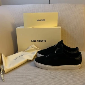 Svarta sneakers från Axel Arigato - Säljer ett par svarta sneakers från Axel Arigato med vit platt sula och dragkedja på ovansidan. Skorna är i mocka och har en stilren design med märkets logga på sidan. Perfekta för dig som gillar minimalistisk stil.