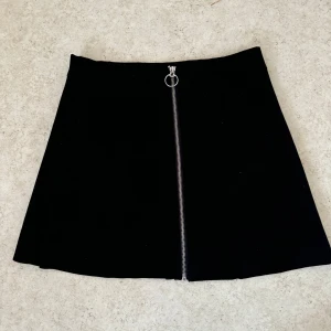 Kjol - Säljer en svart kortkjol från ONLY med en snygg silverfärgad dragkedja framtill. Kjolen har en A-linjeformad siluett och är enkel att matcha med olika toppar. Perfekt för dig som gillar stilrena plagg med en liten detalj.