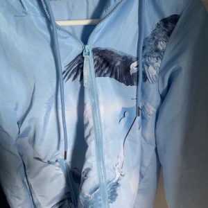 Ljusblå vindjacka med örntryck från Trapstar - Säljer en ljusblå vindjacka från Trapstar med stort örntryck på både fram- och baksida. Jackan har huva, dragkedja och snörning vid halsen. Perfekt för dig som vill sticka ut med en unik design.