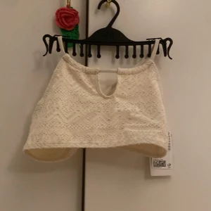 Vit spetsig croppad baddräkt - Säljer en vit croppad baddräkt med spetsdetaljer och ett litet hål framtill. Toppen har smala axelband som korsas i ryggen och ett fint mönster i spets. Perfekt för sommaren och passar till många olika outfits.