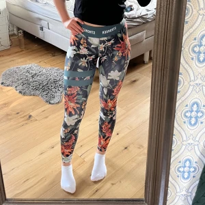 Blommiga träningstights från Stronger - Säljer ett par färgglada träningstights från Stronger i storlek XS. Tightsen har ett blommigt mönster i orange, vitt och grönt med bred resår i midjan🥰🥰