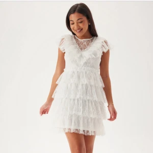 Vit spetsklänning med volanger - Superfin vit klänning i spets med flera lager volanger och v-ringning. Klänningen är ärmlös och har en kort längd, perfekt för sommarens tillfällen. Söt och romantisk stil med fina detaljer. By malina liknande, aldrig använd!