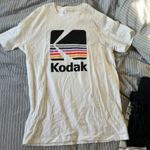 Vit t-shirt med Kodak-logga - Vit t-shirt med stor retro Kodak-logga framtill i svart, rött, orange, gult, lila och rosa. Klassisk passform och rund halsringning. Perfekt för dig som gillar vintage och grafiska tryck.