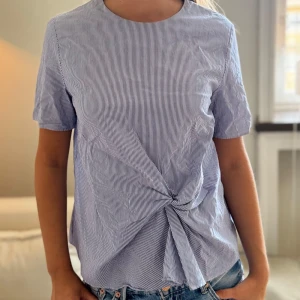 Blårandig blus med knytdetalj - Snygg blårandig blus med korta ärmar och rund hals. Framtill finns en knytdetalj som ger en cool twist till looken. Perfekt till jeans för en avslappnad men stilren outfit.