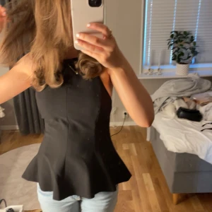 Svart peplumtop från ZARA - Säljer en svart peplumtop från Zara med figurnära passform och volang nertill. Toppen är ärmlös och har en stilren design med rund halsringning och öppen rygg!! 🩷🩷🩷Perfekt att styla med jeans eller kjol.