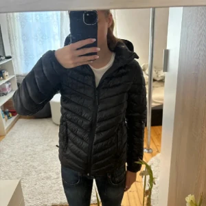 Svart pufferjacka med huva - Säljer en svart pufferjacka med huva och dragkedja framtill. Jackan har lång ärm och en klassisk quiltad design som ger en sportig look. Perfekt för kyligare dagar och enkel att matcha med olika outfits.