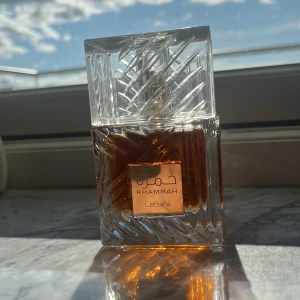 Khamrah Eau de Parfum från Lattafa - Khamrah från Lattafa är en parfym i en lyxig, fyrkantig glasflaska med ett geometriskt mönster och transparent lock. Doften kommer i en elegant svart och guldig förpackning. Flaskan rymmer 100 ml och innehållet har en bärnstensfärgad ton. Ungefär 10 sprays har använts.