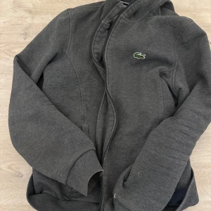 Mörkgrå hoodie från Lacoste Sport - Säljer en mörkgrå hoodie från Lacoste Sport med dragkedja och klassisk krokodillogga på bröstet. Jackan har långa ärmar och är tillverkad i mjukt material som passar perfekt till vardags eller träning. Storlek-S