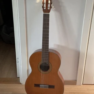 Klassisk akustisk gitarr i trä - Säljer en klassisk akustisk gitarr med naturfärgad träkropp och mörkare hals. Gitarren har sex nylonsträngar och dekorativ rosett runt ljudhålet. Perfekt för både nybörjare och mer erfarna gitarrister som vill ha ett stilrent instrument.