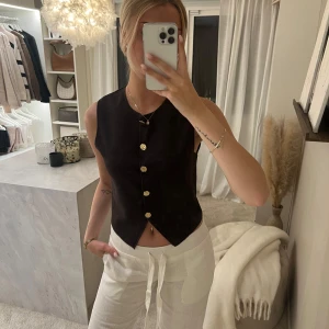 Brun väst med guldknappar från H&M - Stilren brun väst från H&M med snygga guldknappar framtill. Västen har en rak passform och spetsig nederkant. Perfekt att styla över en skjorta eller t-shirt för en trendig look.