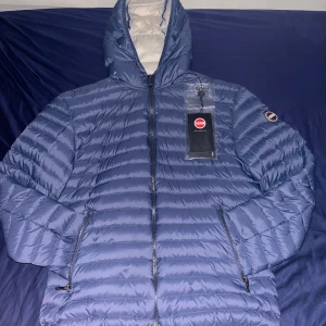 Blå dunjacka från Colmar - Säljer en blå dunjacka från Colmar med huva och dragkedja framtill. Jackan har två sidofickor med dragkedja och quiltad design. Perfekt för kyliga dagar och riktigt snygg passform. Märke på ärmen och innerfoder i ljus färg.