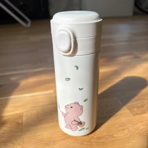 Vit termosmugg med björn motiv - Säljer en söt vit termosmugg med ett tryck av en rosa björn och gröna blad. Muggen har ett praktiskt lock med knapp för enkel öppning och insida i rostfritt stål. Perfekt för att ta med dryck på språng!