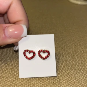 Röda hjärtformade örhängen - Söta örhängen i form av öppna hjärtan, dekorerade med små röda stenar som glittrar. Perfekta för att ge din look en romantisk touch. Metallbas med gnistrande detaljer som verkligen poppar.