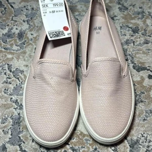 Ljusrosa slip-on sneakers från H&M - Säljer ett par ljusrosa slip-on sneakers från H&M med diskret strukturmönster. Skorna har vit platt sula och rund tå, perfekt för en avslappnad stil. Enkla att ta på tack vare elastiska sidor.