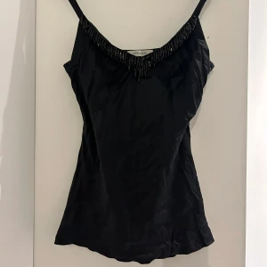 Svart topp med pärldetaljer - Snygg svart linnetopp från Rene Lezard med smala axelband och dekorativa svarta pärlor längs ringningen. Toppen har en figurnära passform och är tillverkad i ett glansigt material som ger en lyxig känsla. Perfekt att styla med jeans eller kjol för en cool look.