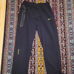 Nike NOCTA svarta mjukisbyxor XL - Svarta mjukisbyxor från Nike NOCTA med gul logga och detaljer. Byxorna har dragsko i midjan, NOCTA-tryck på benet och broderade initialer bak. Sköna och loose fit, perfekta för chill eller streetwear.
