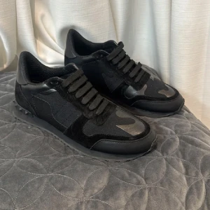 Valentino Rockrunner svarta sneakers - Svarta Valentino Rockrunner sneakers med coola kamouflagemönster i olika svarta nyanser. Skorna har en mix av skinn och mocka, samt klassisk snörning och platt sula. Perfekta för dig som gillar streetstyle och exklusiva detaljer.