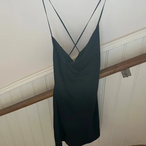 Svart satinklänning med tunna band - Supersnygg svart kortklänning i satin med tunna axelband som korsas i ryggen. Klänningen har en draperad urringning och öppen rygg, perfekt för dig som gillar minimalistisk och elegant stil. Sitter figurnära och känns lyxig på.