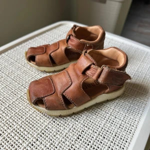 Bruna sandaler i skinn från Bisgaard - Bruna sandaler från Bisgaard i mjukt skinn med vit sula. De har öppen tå, kardborreknäppning och vadderad häl för extra komfort. Perfekta för varma dagar och ger en klassisk look med snygga sömmar och robust känsla.