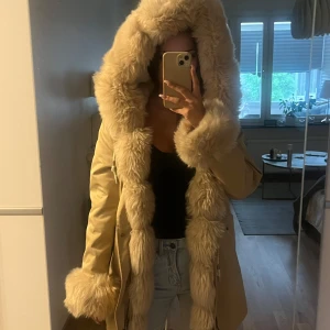 Beige jacka med päls - Säljer en beige parkasjacka med stor fluffig fuskpäls runt huva, ärmslut och längs öppningen. Jackan har dragsko i midjan och stora fickor framtill. Köpt på en webbsida för 3500kr för 2 år sedan.