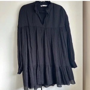 Svart volangklänning från ZARA - Säljer en svart långärmad klänning från ZARA med volangdetaljer och lös passform. Klänningen har v-ringning med liten volangkant och knytband vid halsen. Perfekt för dig som gillar en avslappnad och trendig stil.
