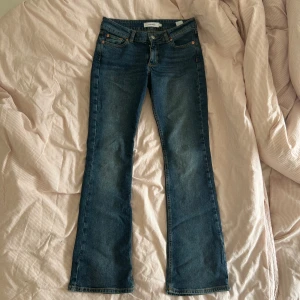 Low waist blå bootcut jeans - Säljer ett par klassiska blå bootcut jeans med fem fickor och orangea sömmar. Jeansen har normal passform och är tillverkade i ett tåligt denimtyg. Perfekta för dig som gillar en tidlös stil med lite utsvängda ben.