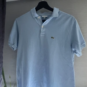 Ljusblå Lacoste piké t-shirt - Snygg ljusblå piké från Lacoste med klassisk krage, två knappar och den ikoniska krokodilloggan på bröstet. Tillverkad i 100% bomull för en skön och luftig känsla. Perfekt för dig som gillar stilren och sportig look. Vid frågor tveka inte att höra av dig😊