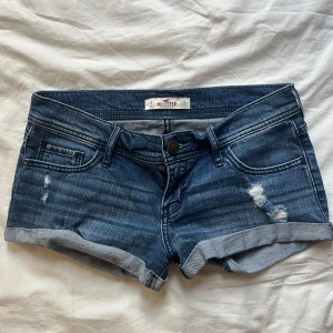 Blå lågmidja jeansshorts från Hollister - Snygga blå lågmidja jeansshorts från Hollister med slitna detaljer framtill och på bakfickan. Storlek w26!