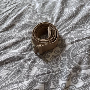 Beige flätat bälte med silverfärgat spänne - Säljer ett stilrent beige flätat bälte med silverfärgat metallspänne. Bältet har detaljer i beige skinn vid spännet och änden. Passar perfekt till jeans eller chinos för en avslappnad look. 105cm