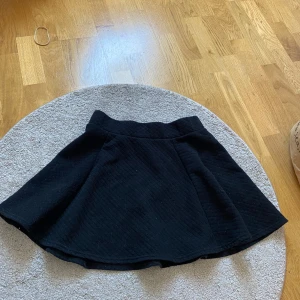 Svart kortkjol från H&M Divided - Svart kortkjol från H&M Divided med  lätt utsvängd form. Kjolen har en synlig dragkedja bak och är tillverkad i ett quiltat material som ger en cool struktur. Perfekt att styla med en enkel topp eller hoodie.