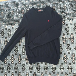 Mörkblå sweatshirt från Morris - Säljer en mörkblå, stickad tröja från Morris i 100% merinoull. Tröjan har rund halsringning, långa ärmar och en liten röd broderad detalj på bröstet. Perfekt för en stilren och enkel look.