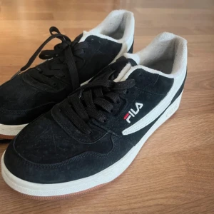 Svarta Fila sneakers i mocka - Snygga svarta Fila sneakers med vit sula och vit detalj längs sidan. Skorna har klassisk Fila-logga i vitt och rött, svarta skosnören och ovandel i mocka. Insidan är fodrad i vitt tyg och yttersulan är i gummi med Fila-mönster. Perfekta för en sportig och stilren look. Använda en gång så ännu i mycket bra skick!