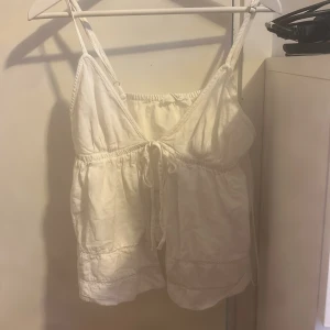 Vit linnetopp med knytband - Säljer en söt vit linnetopp med smala axelband och knytband framtill. Toppen har en luftig passform och är perfekt för varma dagar. Materialet känns lätt och bekvämt mot huden. Den är i storlek S men funkar som M.