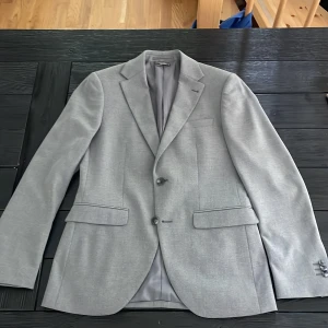 Ljusgrå kavaj Super Slim Fit Riley - Snygg ljusgrå kavaj från Riley i super slim fit-modell. Klassisk design med två knappar framtill, fickor med lock och slät baksida. Fodrad insida med innerficka och diskret märkning. Perfekt för dig som gillar en stilren och modern look.