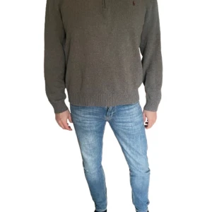 Grå stickad half zip från Polo Ralph Lauren - Snygg grå stickad tröja från Polo Ralph Lauren i bomull. Tröjan har ribbade muddar, hög krage och half zip med läderdetalj på dragkedjan. Klassisk broderad logga på bröstet och långärmad modell. Perfekt för lager-på-lager under höst och vinter.