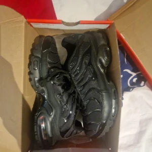 Svarta Nike Air Max Plus sneakers - Säljer ett par svarta Nike Air Max Plus sneakers med klassisk vågig design och synliga luftbubblor i sulan. Skorna har snörning, rund tå och är tillverkade i en kombination av syntet och mesh. Perfekta för dig som gillar streetwear och sportig stil.