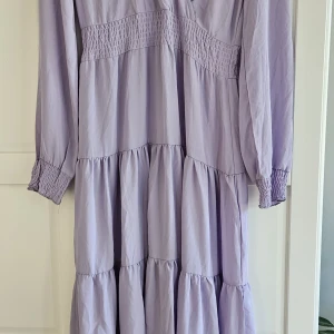 Ljuslila långklänning från Zink - Säljer en romantisk ljuslila långklänning från Zink med v-ringning och smockdetaljer i midjan och vid ärmsluten. Klänningen har långa ärmar och volanger som ger en flowy känsla. Perfekt för sommarens alla tillfällen! Nypris 699