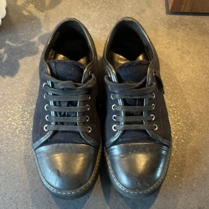 Svarta sneakers från Lanvin i ull och skinn - Snygga svarta sneakers från Lanvin med ovandel i ull och detaljer i skinn. Skorna har rund tå, platt sula och klassisk snörning. Perfekta för dig som gillar stilrena och exklusiva sneakers med en twist. 