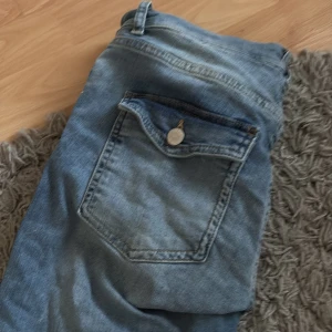 Blå jeans med slitningar - Säljer ett par klassiska blå jeans med slitningar på båda benen och raka ben. Jeansen har normal passform och en bakficka med lock och knapp. Perfekta för en avslappnad stil. Nypris 699kr