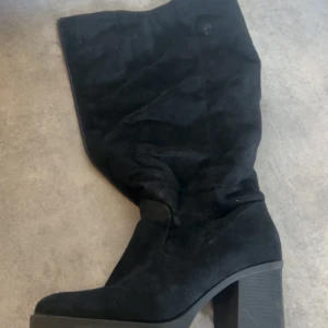 Svarta höga boots med platå - Säljer ett par svarta höga boots med bred klack och platåsula. Skorna har en mjuk mockaliknande yta och en rund tå. Perfekta för att lyfta din outfit med lite extra höjd. Storlek 41 använd av kvinna