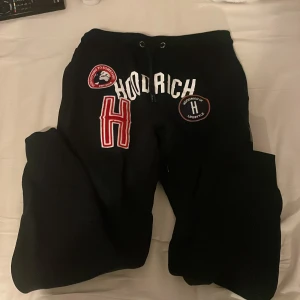 Svarta mjukisbyxor från Hoodrich - Svarta mjukisbyxor från Hoodrich med broderade och tryckta loggor framtill. Byxorna har dragsko i midjan och mjukt material på insidan. Snygga detaljer med patchar och stor röd H på ena benet. Perfekta för en chill och streetwear-look. Finns ett pyttelitet hål bak på byxorna.