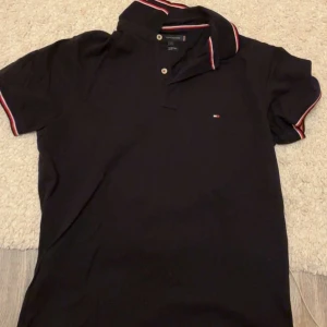 Svart pikétröja Tommy Hilfiger - Svart pikétröja från Tommy Hilfiger i slim fit med klassisk krage och knäppning framtill. Tröjan har röda, vita och blå ränder på krage och ärmslut samt liten logga på bröstet. Tillverkad i ekologisk bomull, perfekt för en clean och stilren look.