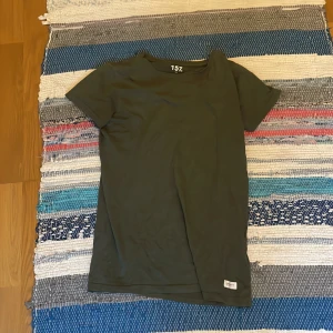 Mörkgrön t-shirt från 157 - En basic mörkgrön t-shirt från 157 med klassisk passform och rund hals. T-shirten är kortärmad och har en liten vit etikett nertill. Perfekt för en clean och enkel stil.