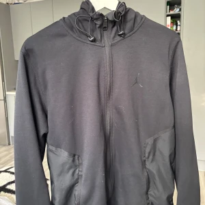 Svart hoodiejacka från Jordan - Säljer en svart hoodiejacka från Jordan med dragkedja och justerbar huva. Jackan har två fickor framtill och en diskret Jumpman-logga på bröstet. Perfekt för en sportig och avslappnad stil.