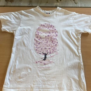 Vit t-shirt med rosa trädtryck från BAPE - Vit t-shirt från BAPE med ett stort tryck av ett rosa blommande träd på framsidan. Klassisk passform och korta ärmar. Ikonisk BAPE-logga på ärmens etikett. Perfekt för dig som gillar streetwear och unika prints.