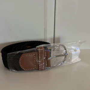 Svart flätat bälte  - Snyggt svart flätat bälte med bruna skinn-detaljer och silverspänne. Perfekt accessoar för att lyfta din outfit. Bältet har klassisk design och passar till många olika stilar. Är elastisk!