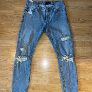 Blå slitna jeans med hål - Säljer ett par blå jeans ifrån Tiger of Sweden med flera slitningar och hål på både fram- och baksidan, särskilt vid knäna och bakfickorna. Klassisk femficksmodell rak passform. Perfekta för en avslappnad och trendig stil.