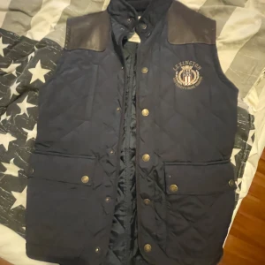 Mörkblå quiltad dunväst Lexington S - Snygg mörkblå quiltad dunväst från Lexington med skinnliknande axelpartier och tryckknappar framtill. Västen har fyra stora fickor med lock och broderad logga på bröstet. Perfekt för lager-på-lager och klassisk stilren stil. Strl S 