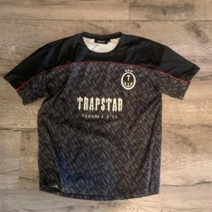 Trapstar jersey - Snygg svart och grå t-shirt från Trapstar med korta ärmar, röd detaljsöm och coolt mönster över hela framsidan. Stort Trapstar-tryck och emblem framtill, samt siffran 22 och Trapstar-tryck på ryggen. Perfekt för dig som gillar streetwear.